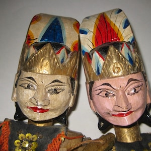 May include: Two wooden puppets with painted faces and ornate headdresses. The puppets have gold crowns and wear black and gold clothing. The headdresses are cream-colored with blue, red, and yellow painted designs. The puppets' faces are painted with red lips and black eyebrows.