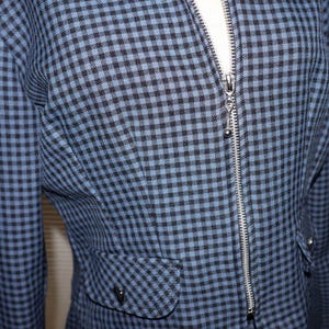 May include: A close-up of a blue and black checkered jacket with a silver zipper. The jacket has two pockets and a small silver charm hanging from the zipper. The fabric has a textured appearance.