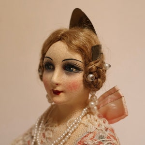 May include: A vintage doll with a painted face, blue eyes, and red lips. The doll has blonde hair styled with pearl accents and wears a pearl necklace. The doll is dressed in a cream-colored dress with lace and a pink bow.