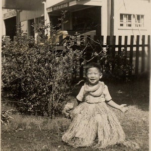 May include: A black and white vintage photograph of a smiling child wearing a light-colored short-sleeved shirt and a textured grass skirt with a matching lei around the neck, standing outdoors in front of a building with signs reading SERVICE.
