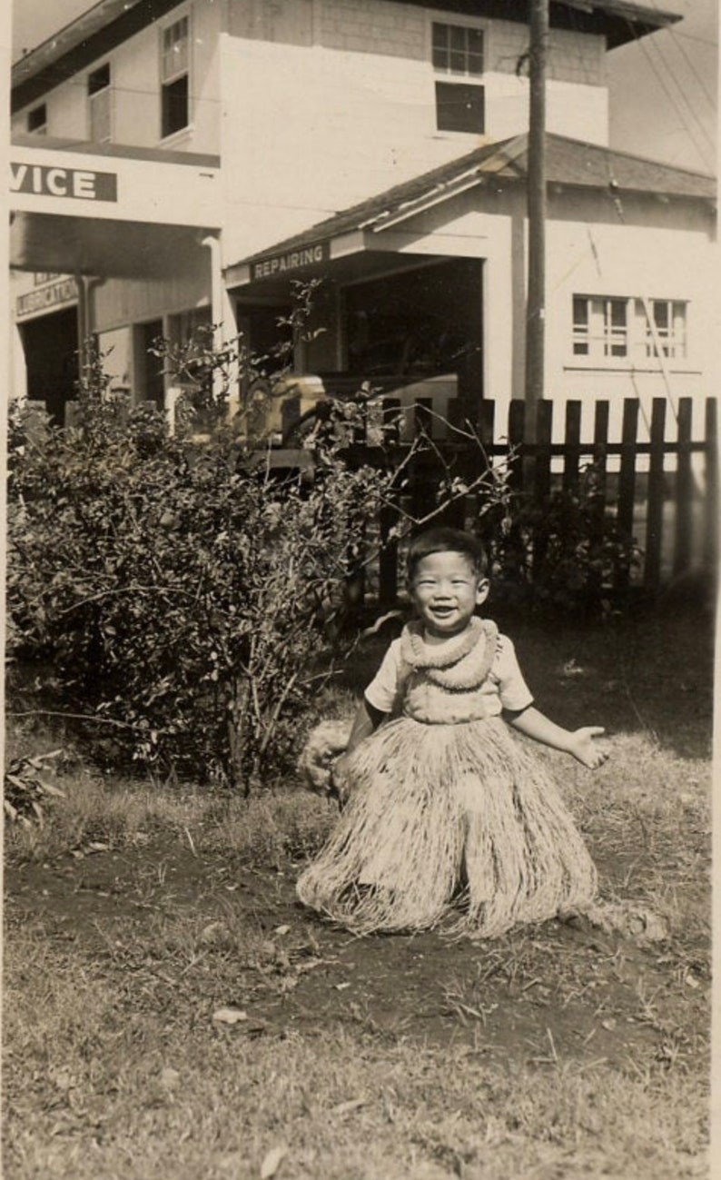 May include: A black and white vintage photograph of a smiling child wearing a light-colored short-sleeved shirt and a textured grass skirt with a matching lei around the neck, standing outdoors in front of a building with signs reading SERVICE.