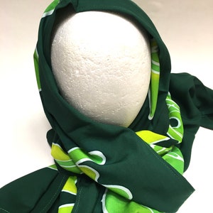 May include: A dark green scarf with a repeating pattern of white and lime green abstract shapes. The scarf is draped over a white mannequin head, showcasing the fabric's drape and design. The scarf's edges are neatly finished.