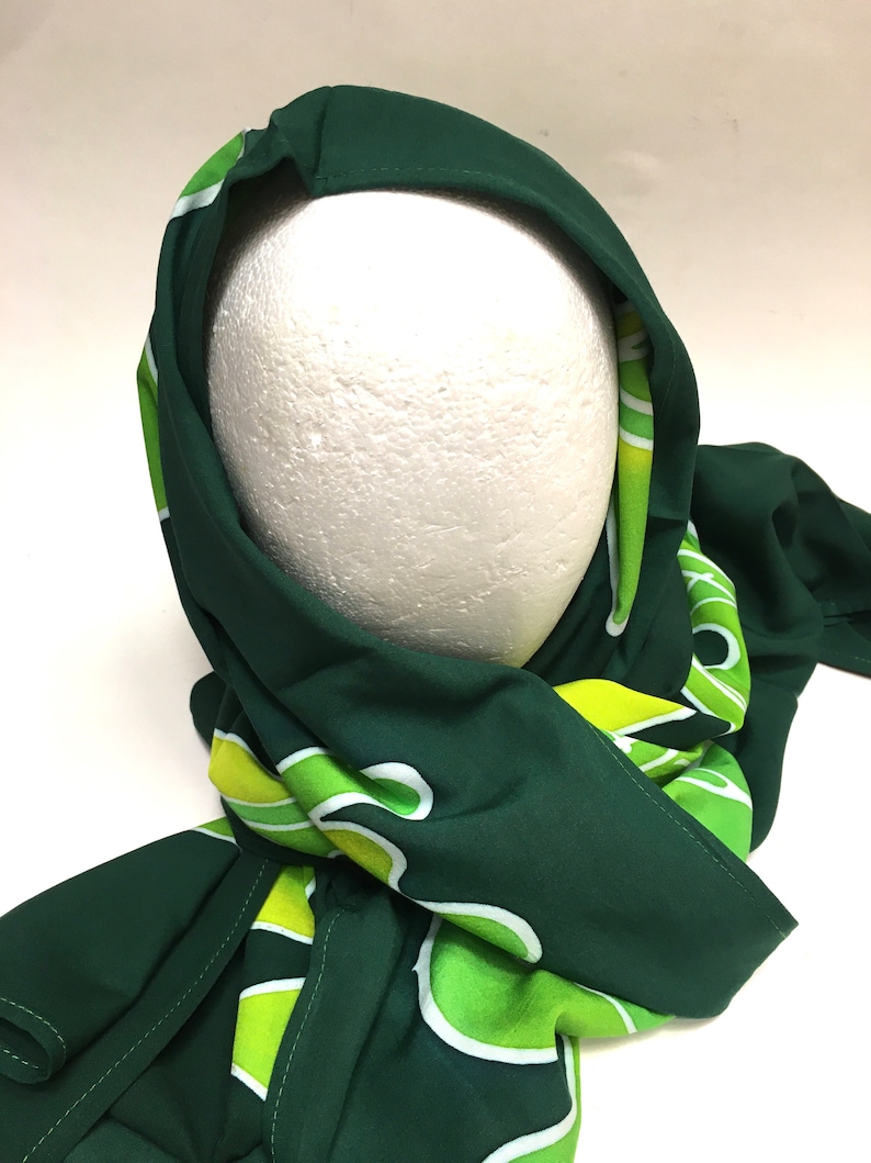 May include: A dark green scarf with a repeating pattern of white and lime green abstract shapes. The scarf is draped over a white mannequin head, showcasing the fabric's drape and design. The scarf's edges are neatly finished.