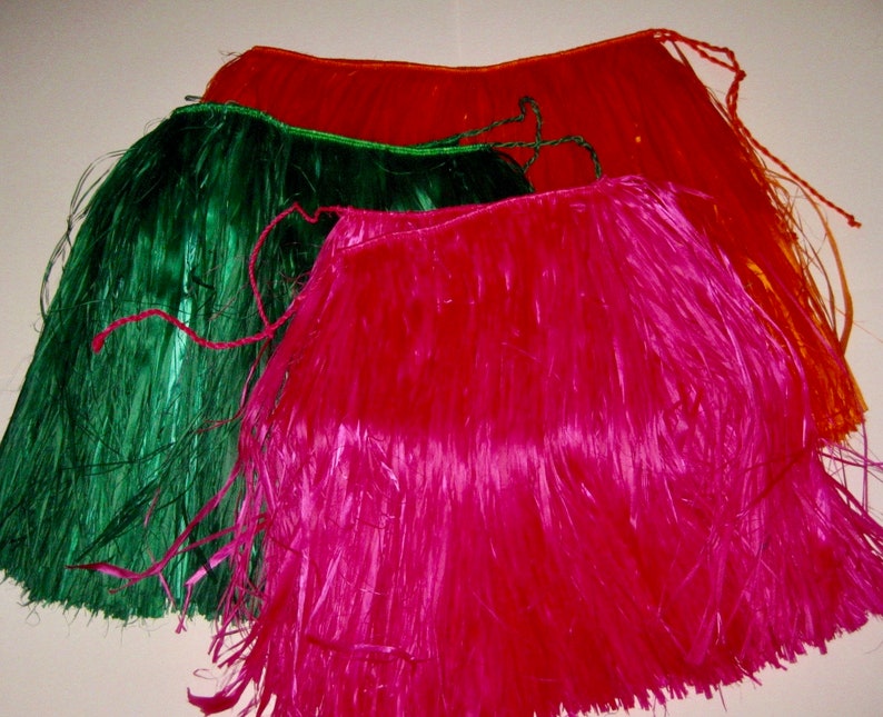 May include: Three hula skirts in bright colors, including a vibrant pink, a deep forest green, and a warm orange. Each skirt is constructed from thin, glossy synthetic strips attached to a braided waistband with long ties for securing.