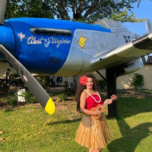 May include: A person wearing a red strapless top, a tan raffia hula skirt, and a red hibiscus flower in their hair, holding a wooden ukulele. The person stands in front of a blue and silver vintage propeller airplane with the text The West by Gowd Virginian.