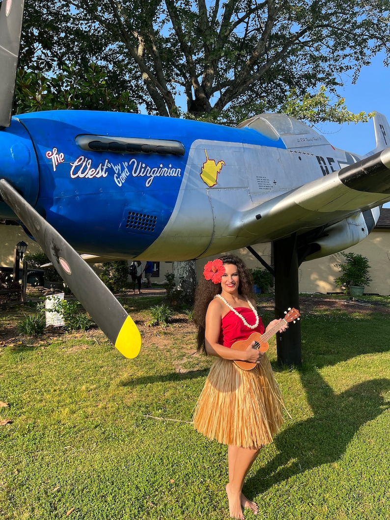 May include: A person wearing a red strapless top, a tan raffia hula skirt, and a red hibiscus flower in their hair, holding a wooden ukulele. The person stands in front of a blue and silver vintage propeller airplane with the text The West by Gowd Virginian.