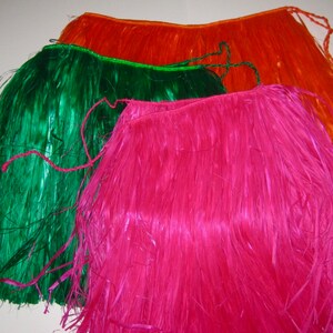 May include: Three hula skirts in bright orange, vibrant green, and hot pink. Each skirt is constructed from long, thin, synthetic plastic strips attached to a braided waistband with matching tie closures at the ends for securing the garment.