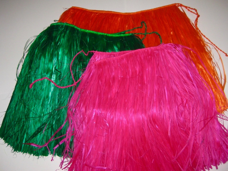 May include: Three hula skirts in bright orange, vibrant green, and hot pink. Each skirt is constructed from long, thin, synthetic plastic strips attached to a braided waistband with matching tie closures at the ends for securing the garment.
