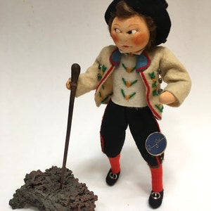 May include: A vintage doll dressed in traditional attire, holding a wooden walking stick. The doll wears a cream-colored jacket with colorful embroidery, black pants, red socks, and black shoes. A dark hat sits atop the doll's head.