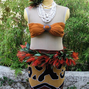 May include: A brown strapless top with a wooden buckle, a white shell necklace, and a skirt with a brown and black pattern. The skirt has a decorative belt of red and green feathers. A flower hairpiece completes the ensemble.