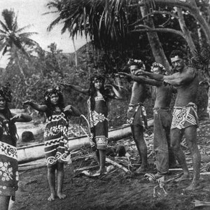 May include: Black and white photograph of a group of people in traditional attire, possibly performing a dance. The individuals are adorned with floral headpieces and patterned wraps, set against a tropical backdrop of palm trees and water.