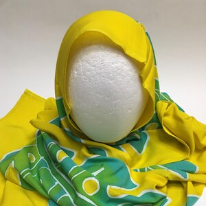 May include: A yellow head covering with a green and teal pattern. The head covering is draped over a white foam head. The fabric has a bright, cheerful appearance.