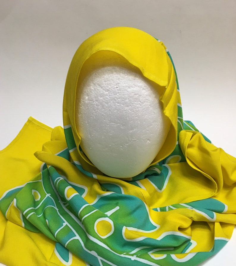May include: A yellow head covering with a green and teal pattern. The head covering is draped over a white foam head. The fabric has a bright, cheerful appearance.