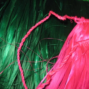 May include: A bright pink hula skirt featuring a twisted cord waistband and long, thin, fibrous strands. The skirt is positioned against a background of deep green, long, thin, fibrous strands that contrast with the vibrant pink color.