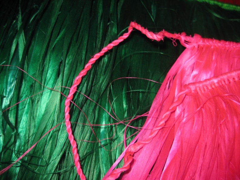 May include: A bright pink hula skirt featuring a twisted cord waistband and long, thin, fibrous strands. The skirt is positioned against a background of deep green, long, thin, fibrous strands that contrast with the vibrant pink color.