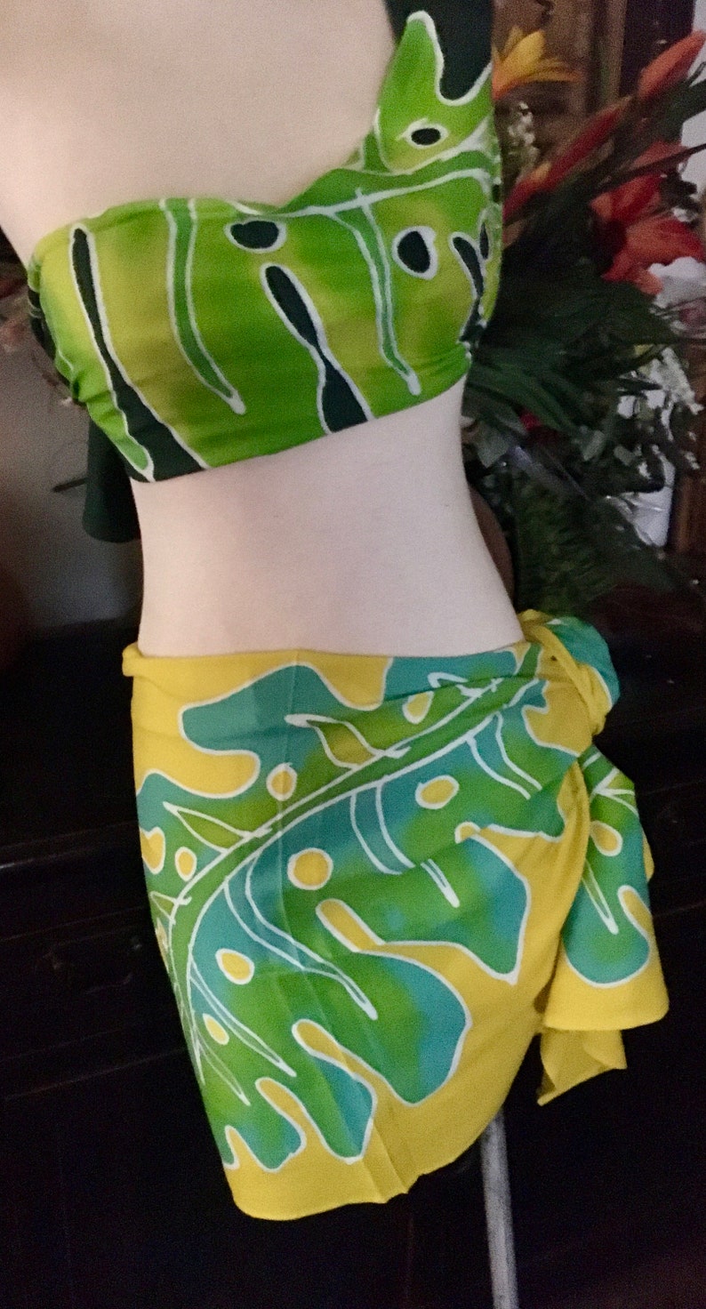 May include: A two-piece swimsuit set featuring a green, yellow, and teal leaf pattern. The top is a bandeau style, and the sarong-style bottom is tied at the waist. The fabric has a tropical, summery vibe.