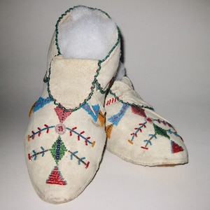 May include: A pair of off-white moccasins with colorful beadwork. The beaded designs include geometric patterns in red, blue, green, and yellow. The edges are trimmed with green beads.