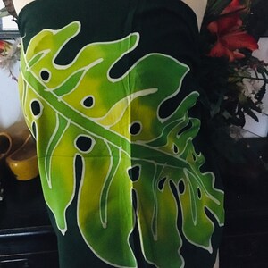 May include: Dark green sarong with a large, stylized leaf design in shades of green and yellow. The leaf has black accents and white outlines. The sarong is draped on a mannequin, showcasing the fabric's pattern and drape.