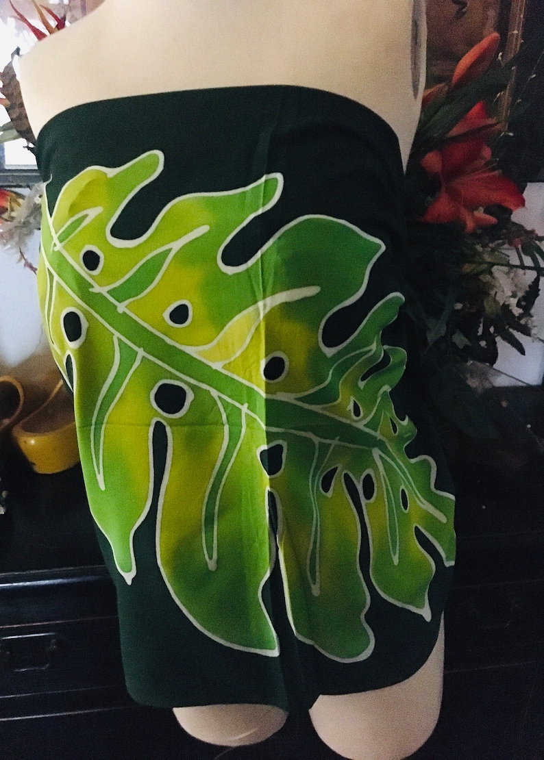 May include: Dark green sarong with a large, stylized leaf design in shades of green and yellow. The leaf has black accents and white outlines. The sarong is draped on a mannequin, showcasing the fabric's pattern and drape.