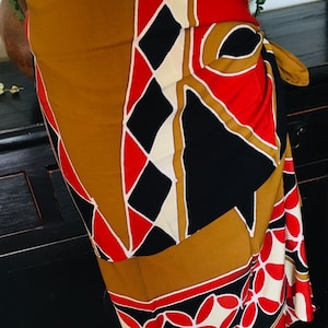 May include: A wrap skirt with a geometric pattern in shades of brown, red, black, and white. The skirt features a repeating floral design along the bottom edge and a tie closure.