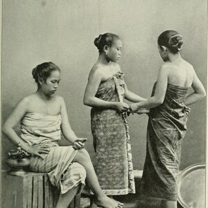 May include: Three women in traditional attire, captured in a sepia-toned photograph. One woman sits on a wooden crate, while the other two stand, possibly adjusting a woven item. The women are wearing patterned wraps.