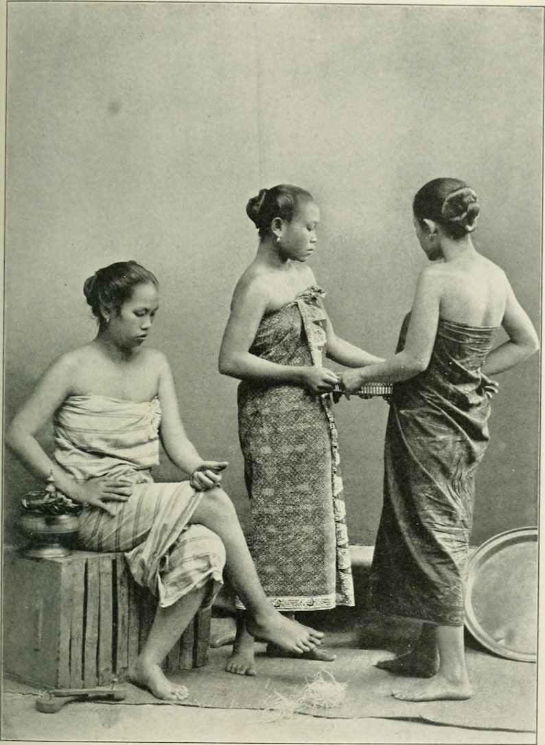 May include: Three women in traditional attire, captured in a sepia-toned photograph. One woman sits on a wooden crate, while the other two stand, possibly adjusting a woven item. The women are wearing patterned wraps.