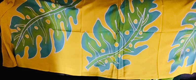 May include: Yellow fabric with a repeating pattern of large tropical leaves. The leaves are green and blue with white outlines, set against a bright yellow background. The design is likely for home decor or apparel.