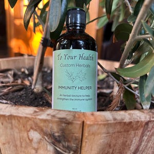 Puede incluir: Una botella de vidrio marrón con una tapa negra, etiquetada con "To Your Health Custom Herbals" y "IMMUNITY HELPER". La etiqueta también dice "Una tintura herbal para ayudar a fortalecer el sistema inmunológico" y "100 ml".