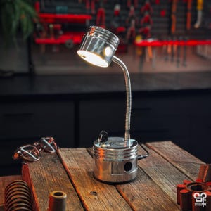 May include: A silver metal desk lamp constructed from an engine piston base and a flexible gooseneck arm. The lamp features a cylindrical piston head as the light housing, a black ignition key switch on the base, and a power cord extending from the side.