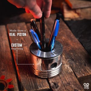 Könnte beinhalten: Ein zylindrischer Stiftehalter aus Metall, gefertigt aus einem echten Motor-Kolben mit individueller Gravur am oberen Rand. Der silberfarbene Halter enthält drei Stifte in Schwarz und Blau. Der Text lautet: Made from a REAL PISTON + CUSTOM Engraving.