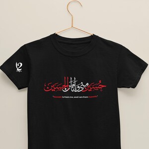 K&ouml;nnte beinhalten: Schwarzes T-Shirt mit arabischer Kalligraphie in Rot und Wei&szlig;, mit dem Satz "Hussein ist von mir, und ich bin von Hussein." Ein wei&szlig;es Logo befindet sich auf dem linken &Auml;rmel. Das T-Shirt h&auml;ngt an einem goldenen Kleiderb&uuml;gel.