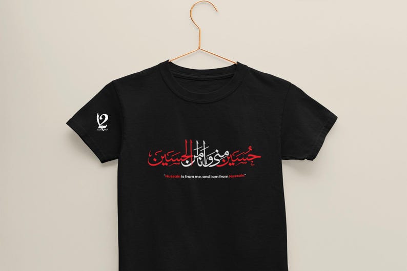 K&ouml;nnte beinhalten: Schwarzes T-Shirt mit arabischer Kalligraphie in Rot und Wei&szlig;, mit dem Satz "Hussein ist von mir, und ich bin von Hussein." Ein wei&szlig;es Logo befindet sich auf dem linken &Auml;rmel. Das T-Shirt h&auml;ngt an einem goldenen Kleiderb&uuml;gel.