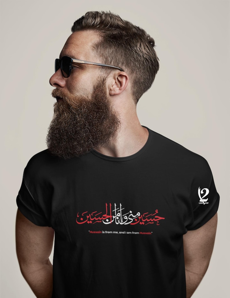 K&ouml;nnte beinhalten: Schwarzes T-Shirt mit roter und wei&szlig;er arabischer Kalligraphie und dem englischen Text "Hussain is from me, and I am from Hussain." Das Shirt hat einen Rundhalsausschnitt und kurze &Auml;rmel. Ein Logo ist auf der rechten Schulter sichtbar.