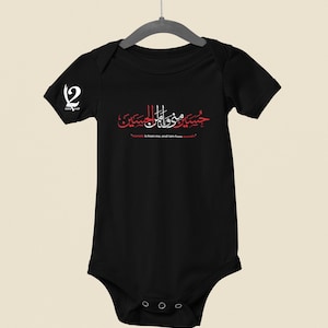 K&ouml;nnte beinhalten: Schwarzer Baby-Body mit roter arabischer Kalligraphie und dem Text "Hussain ist von mir, und ich bin von Hussain." Auf dem linken &Auml;rmel befindet sich ein wei&szlig;es Logo. Der Body h&auml;ngt an einem Metallb&uuml;gel.