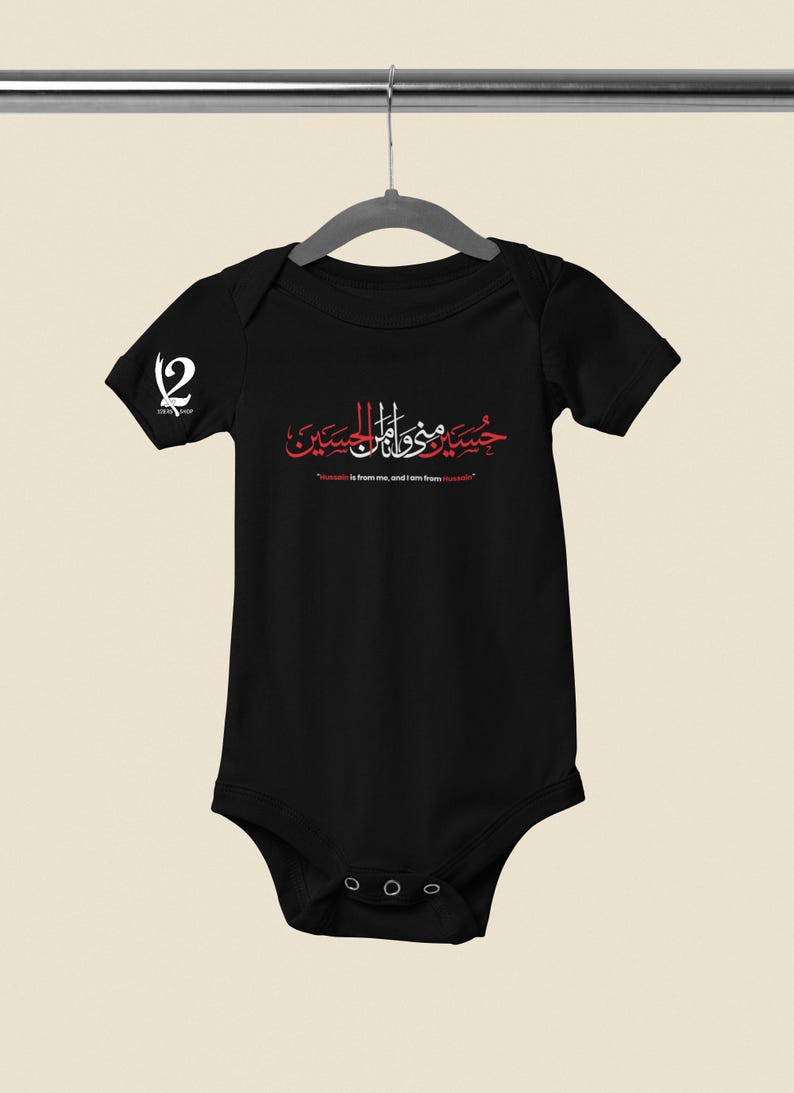 K&ouml;nnte beinhalten: Schwarzer Baby-Body mit roter arabischer Kalligraphie und dem Text "Hussain ist von mir, und ich bin von Hussain." Auf dem linken &Auml;rmel befindet sich ein wei&szlig;es Logo. Der Body h&auml;ngt an einem Metallb&uuml;gel.