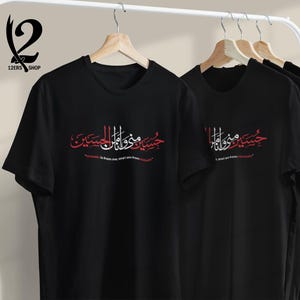 K&ouml;nnte beinhalten: Schwarze T-Shirts mit arabischer Kalligraphie in Rot und Wei&szlig;, die auf einem Gestell h&auml;ngen. Der Text ist mittig auf der Vorderseite der Shirts angeordnet. Die Shirts werden auf Holzb&uuml;geln vor einem neutralen Hintergrund pr&auml;sentiert.