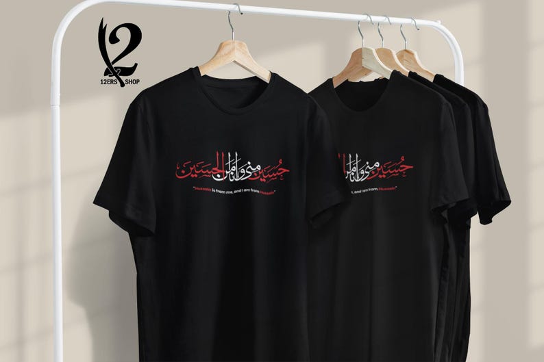 K&ouml;nnte beinhalten: Schwarze T-Shirts mit arabischer Kalligraphie in Rot und Wei&szlig;, die auf einem Gestell h&auml;ngen. Der Text ist mittig auf der Vorderseite der Shirts angeordnet. Die Shirts werden auf Holzb&uuml;geln vor einem neutralen Hintergrund pr&auml;sentiert.