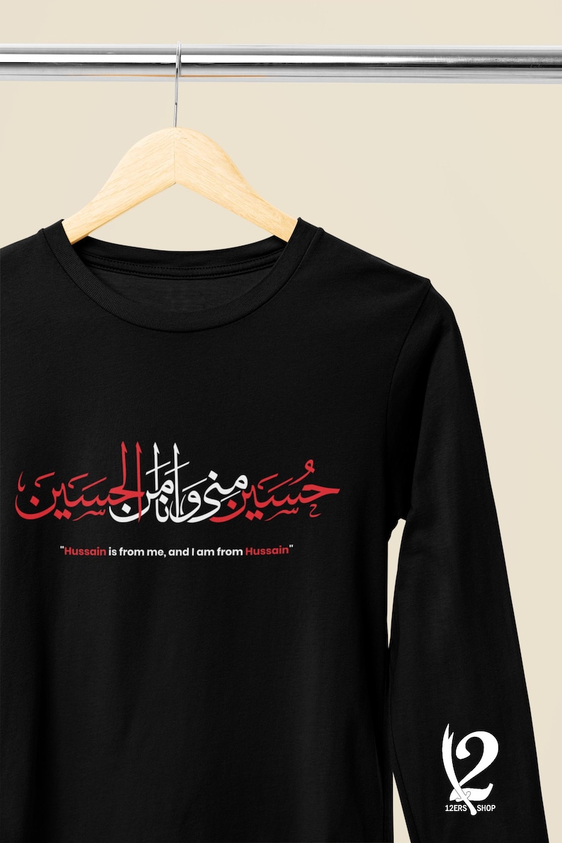 K&ouml;nnte beinhalten: Schwarzes Langarmshirt mit roter und wei&szlig;er arabischer Kalligraphie und dem englischen Text "Hussain is from me, and I am from Hussain." Das Shirt hat einen Rundhalsausschnitt und ein Logo auf dem &Auml;rmel.