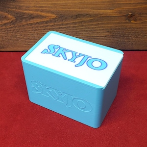 以下が含まれることがあります： 上面と側面に青い文字で「SKYJO」と書かれた水色の長方形の箱。箱は赤い表面に置かれ、背景は木です。
