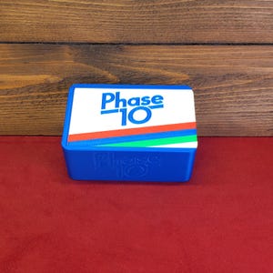 May include: A blue Phase 10 card game box with a white lid. The lid features the game's name in blue, with red, green, and blue stripes. The game's name is also printed on the box's side.