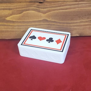 May include: A white rectangular plastic box featuring a playing card design on the lid with black spade and club symbols and red heart and diamond symbols. The front side of the box displays embossed spade, diamond, club, and heart shapes.
