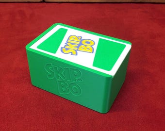 May include: A green box of the card game Skip-Bo, with the game's logo on the top and side. The logo is yellow and blue on a white background. The box is on a red surface.