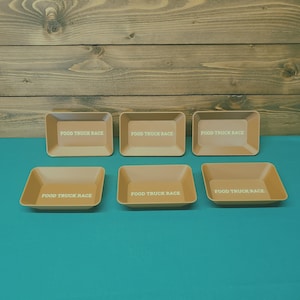 May include: Six rectangular, light brown serving trays with the text "FOOD TRUCK RACE" in white. The trays are arranged on a teal surface against a wooden background.
