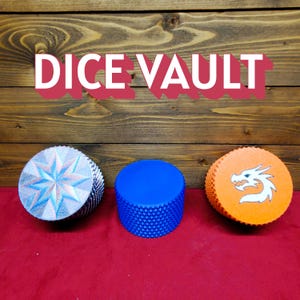 Puede incluir: Tres cajas cilíndricas para dados. Una es plateada con un diseño de estrella, otra es azul y la tercera es naranja con un gráfico de dragón blanco. El texto "DICE VAULT" está encima de las cajas. Las cajas están sobre una superficie roja con un fondo de madera.