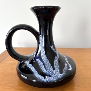 May include: A dark blue ceramic vase with a handle. The vase has a bulbous base and a flared opening. White and blue abstract patterns decorate the surface. The vase is on a wooden surface.