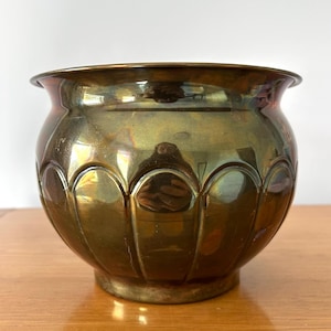 Peut inclure: Un pot de fleurs en laiton vintage avec un corps arrondi et des détails festonnés. Le pot a un bord large et légèrement évasé et une teinte dorée chaude. Il repose sur une petite base circulaire. La surface reflète la lumière.