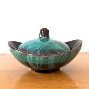 May include: A vintage ceramic bowl with a lid. The bowl is a deep teal colour with brown accents and a brown lid handle. The bowl has a unique shape with flared sides and a glossy finish. The bowl is likely used for serving or display.