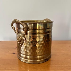 Könnte beinhalten: Vintage Messing-Eimer mit gehammerter Textur und Löwenkopf-Details. Der Eimer hat zwei Griffe mit Seil-Design. Der zylindrische Eimer steht auf einer Holzoberfläche.