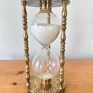 May include: An hourglass with a brass frame and clear glass bulbs. The top bulb is filled with white sand, and the bottom bulb is empty. The base and top have Roman numerals. The hourglass is a decorative item.
