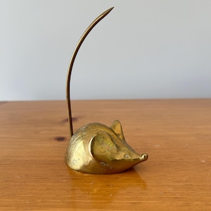 May include: A brass mouse ornament with a curved tail. The mouse is in a sitting position with its ears perked up. The ornament is made of solid brass and has a shiny, golden finish. The mouse is sitting on a wooden surface.